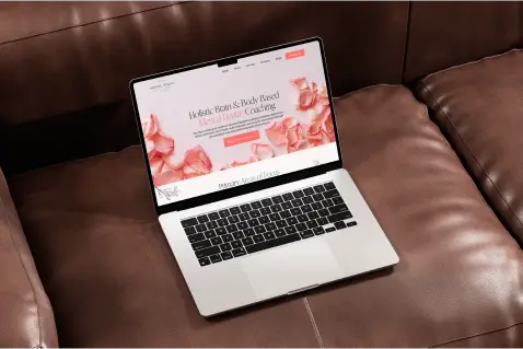 Laptop displaying a modern wellness coaching website design featuring soft pink tones, positioned on a leather couch, emphasizing digital solutions for therapists and wellness coaches.